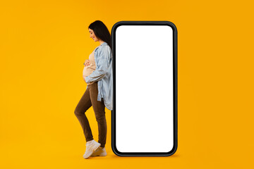 Happy pregnant woman leaning on huge blank cellphone and hugging her belly, advertising mobile app or website, mockup