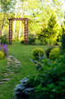 Beautiful summer garden view with curvy stone pathway and wooden archway. Natural woodland cottage garden with hostas, conifers and shrubs