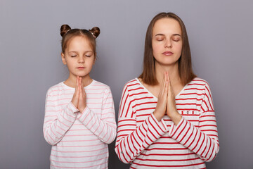 Portrait of calm relaxed mother and daughter wearing casual clothes standing isolated over gray background, keep palms together, praying or practicing yoga.