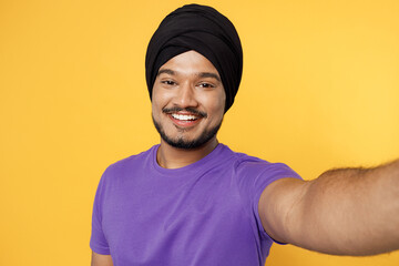 Wall Mural - Close up smiling happy fun devotee Sikh Indian man ties his traditional turban dastar wear purple t-shirt do selfie shot pov on mobile cell phone isolated on plain yellow background studio portrait