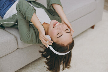 Young woman of Asian ethnicity wearing casual clothes headphones listen to music close eyes lay down on grey sofa couch stay at home hotel flat rest relax spend free spare time in living room indoor.