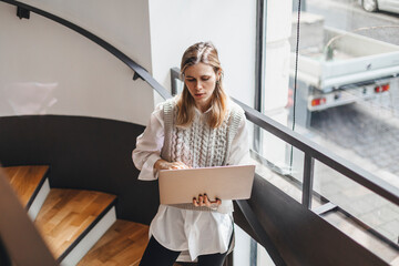 Serious young woman standing on the stairs typing on her laptop. Attractive young blonde business lady work at home, office or cafe with calm face. Girl wear knitted vest and white shirt.