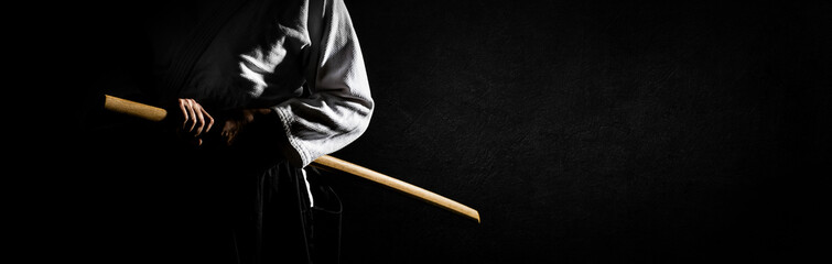 Wall Mural - A person in black hakama standing in fighting pose with wooden sword bokken over grunge dark background banner. Shallow depth of field. SDF. 