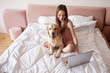 © Богдан Маліцький - girl lies on bed with golden retriever dog and uses laptop, female freelancer with pet looks at the computer