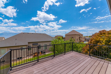 Terrace of a house overlooking houses in the neighborhood under bright blue sky. The deck has a wooden floor and safety railings with view of trees and grassy lawn.