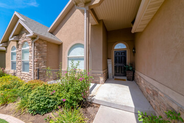 Sticker - Entrance of a house with painted light brown stucco and stone veneer wall. House with plants near the arched windows on the left and black front door with transom and panel and plants on the right.