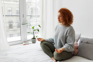 Fototapete - Young plus-size redhead woman with fluffy curly hair sitting in lotus posture on bed near big window keeping hands on knees, meditating, trying to relax her mind and relieve stress