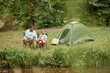 © Seventyfour - Front view at black mother and daughter camping together outdoors and fishing by river, copy space
