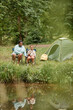 © Seventyfour - Vertical portrait of black mother and daughter camping together outdoors and fishing by river