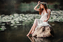 Beautiful Red Haired Girl In White Vintage Dress And Wreath Of Flowers Sitting On The Shore Of River. Fairytale Story. Warm Art Work.