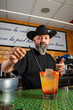 © Rafa - hombre barman con gorro de vaquero preparando un cóctel azul en la barra del bar