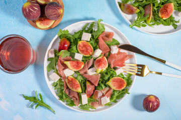 Autumn figs salad with arugula, feta, raw ham in white plate on light blue table background served with rose wine. Flexitarian diet, paleo diet fall or summer salad, close up, top view, food flat lay