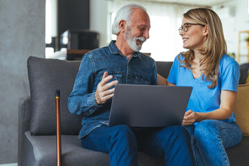 Wall Mural - Friendly pretty young woman nurse assisting disabled senior man at home using laptop, sitting at desk in front of computer, having conversation and smiling, man touching gadget screen