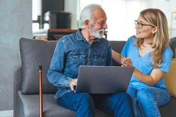 Wall Mural - Senior man with disability learning to pay bills online with laptop together with social worker. Nurse help senior retirement man working with laptop. Active senior man using laptop with female carer