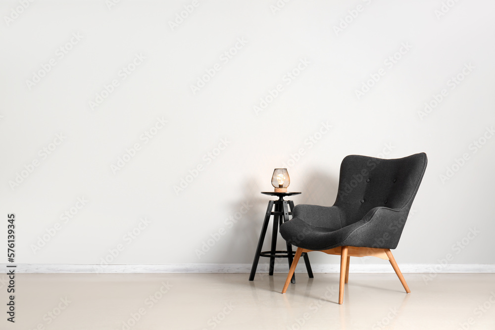 Stylish grey armchair and end table with glowing lamp near white wall