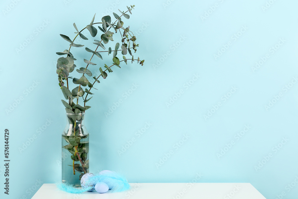 Vase with eucalyptus branches and Easter eggs on table near blue wall