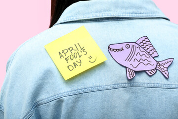 Wall Mural - Sticky paper with text APRIL FOOL'S DAY and fish on woman's back against pink background, closeup