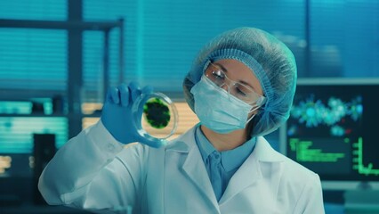 Wall Mural - A female scientist examines a petri dish with a spotted green sample. A woman in a white medical gown, mask, blue gloves, goggles and a cap examines the spores of a virus or bacteria. Close up.