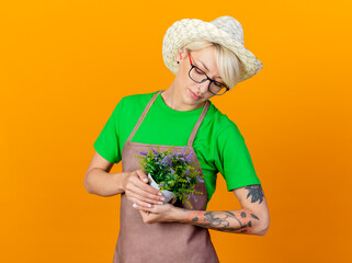 Wall Mural - young gardener woman with short hair in apron and hat holding potted plant looking at it with love standing over orange background