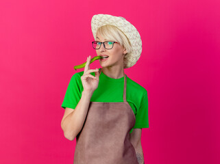 Wall Mural - young gardener woman with short hair in apron and hat holding green chili pepper using it like cigar looking confident standing over pink background