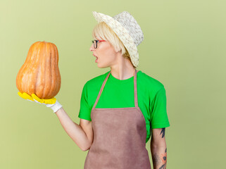 Wall Mural - young gardener woman with short hair in apron and hat wearing rubber gloves holding pumpkin looking at it surprised standing over light background