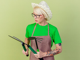 Wall Mural - young gardener woman with short hair in apron and hat holding hedge clippers looking at it intrigued standing over light background