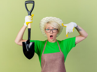 Wall Mural - young gardener woman with short hair in apron and hat wearing rubber gloves holding shovel pointing with index finger looking surprised standing over light background