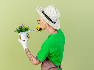 Wall Mural - young gardener woman with short hair in apron and hat holding potted plant measuring it with measure tape looking confident standing over light background