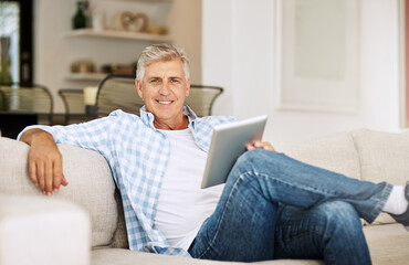 Poster - I always liked keeping up with the latest news.... Shot of a mature man using his digital tablet while relaxing on his sofa at home.