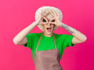 Wall Mural - young gardener woman with short hair in apron and hat looking at camera through fingers making binocular gesture with fingers standing over pink background