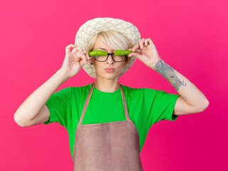 Wall Mural - young gardener woman with short hair in apron and hat holding halves of green chili pepper over her closed eyes standing over pink background