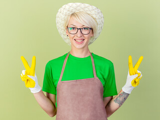 Wall Mural - young gardener woman with short hair in apron and hat wearing rubber smiling showing v-sign standing over light background