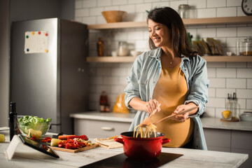 Beautiful pregnant woman preparing delicious food. Smiling woman cooking pasta at home.