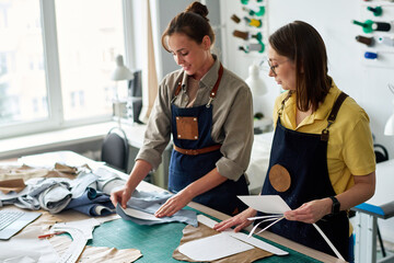 Young tailor holding sewing pattern on piece of blue leather while working with her colleague over new clothing item for fashion collection