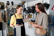 © AnnaStills - Happy young female customer looking at shop assistant offering her new handmade leather items while choosing creative stuff to buy