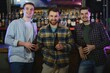© Serhii - Three young men in casual clothes are smiling, holding bottles of beer while standing near bar counter in pub