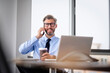 © gzorgz - Mid aged businessman sitting at office desk and using cellphone and laptop