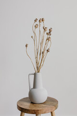 a ceramic vase on a wooden stool with dried poppy heads on branch. minimal home decoration, copy spa