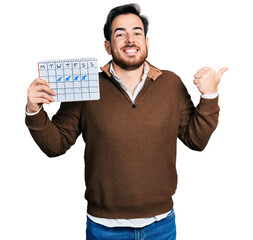 Young hispanic man holding travel calendar pointing thumb up to the side smiling happy with open mouth