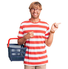 Sticker - Handsome caucasian man with beard holding supermarket shopping basket smiling happy pointing with hand and finger to the side