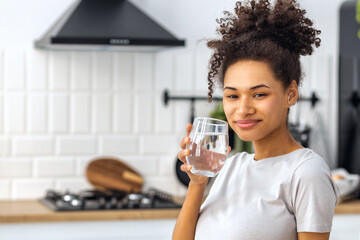 Portrait of African American young woman holding  glass of water standing in the kitchen at home looking at the camera and smiling, wellness healthy food concept