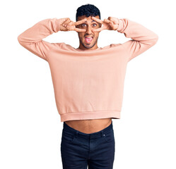 Wall Mural - Young hispanic man wearing casual clothes doing peace symbol with fingers over face, smiling cheerful showing victory