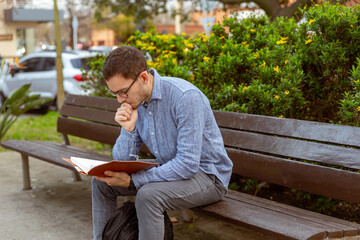 hombre joven, caucásico, de 25 años, leyendo un libro, en un banco de un parque.