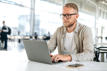 Concentrated busy caucasian man with glasses, in stylish casual wear, IT specialist, software tester, programmer, digital developer, works in a laptop in modern office, testing a new program