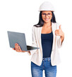 © Krakenimages.com - Young beautiful latin girl wearing architect hardhat holding laptop smiling happy and positive, thumb up doing excellent and approval sign