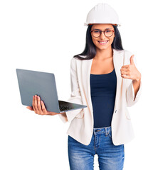 Wall Mural - Young beautiful latin girl wearing architect hardhat holding laptop smiling happy and positive, thumb up doing excellent and approval sign