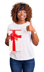 Wall Mural - Young african american woman holding gift smiling happy and positive, thumb up doing excellent and approval sign