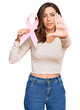 © Krakenimages.com - Young brunette woman holding pink cancer ribbon with open hand doing stop sign with serious and confident expression, defense gesture