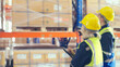 © Monster Ztudio - Warehouse workers holding digital tablet checking inventory management packaging boxes. Two staff wearing vest and safety helmet and walking count the box goods on shelves at storehouse