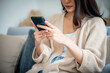 © Monster Ztudio - Happy young asian woman relax on comfortable couch at home texting messaging on smartphone, smiling girl use cellphone chatting, browse wireless internet on gadget, shopping online from home